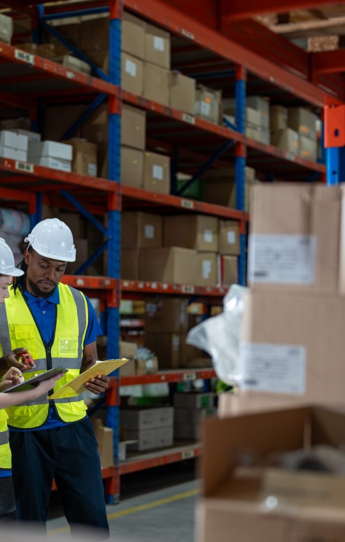 Professional female and man warehouse worker with digital tablet talking together and checking stock products in boxes on shelves at warehouse factory store. Logistics, Distribution Center concept.