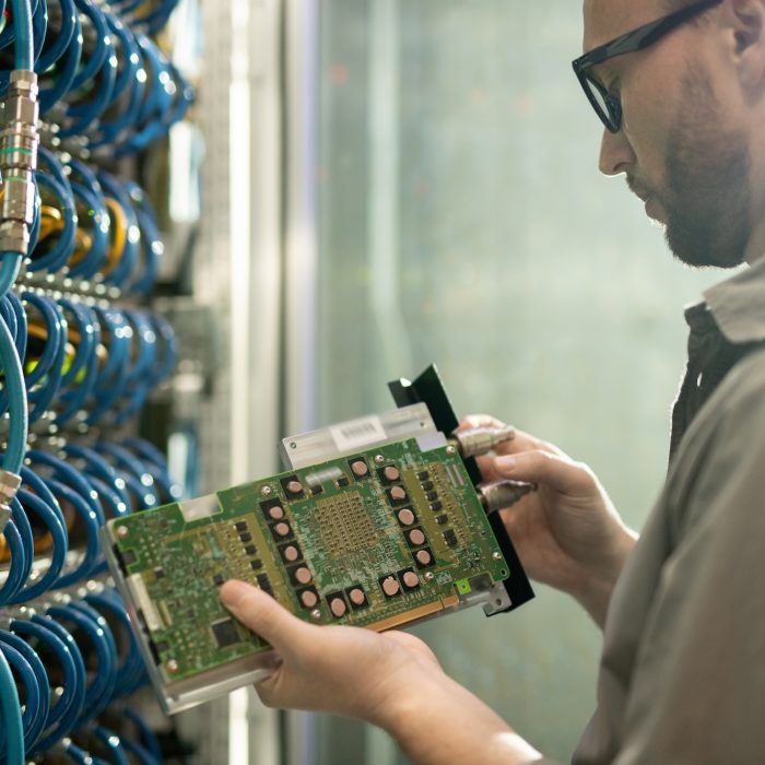 Serious skilled young IT engineer standing at server of database storage and inspecting supercomputer motherboard